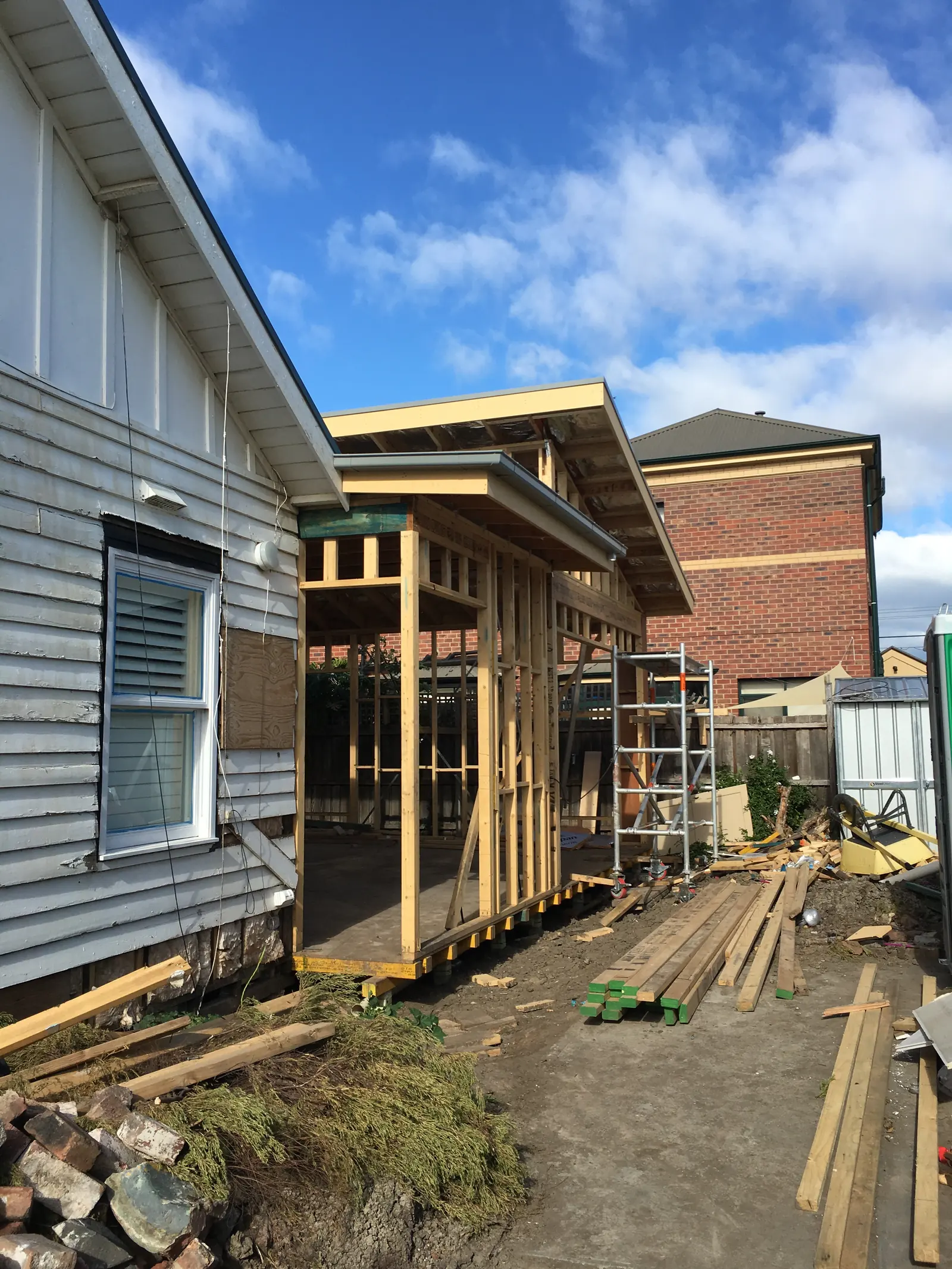 Timber frame of the new rear extension standing beside the original weatherboard house, with scaffolding and stacked timber